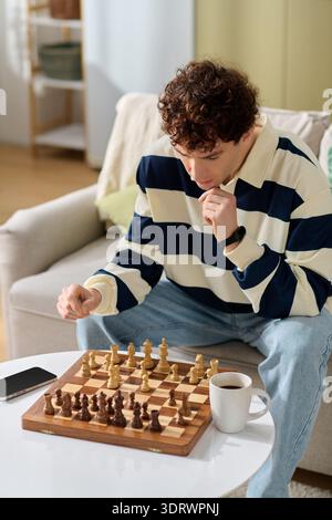 A handsome young man thoughtfully plays chess at home, savoring a warm cup of coffee. Stock Photo