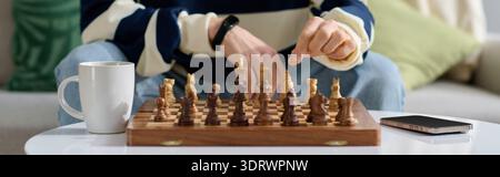 A young man thoughtfully plays chess at home, sipping a warm drink and focused on his strategy. Stock Photo