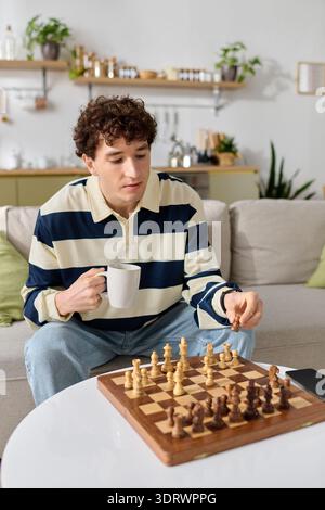 A young man thoughtfully plays chess at home, holding a warm cup of coffee in his hand. Stock Photo