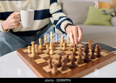 Young man skillfully plays chess on a sunny afternoon, holding a warm cup of coffee. Stock Photo