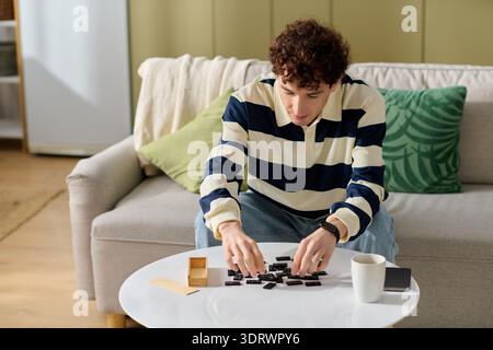 Handsome young man sits at a table, focused on arranging black dominoes in a welcoming space. Stock Photo