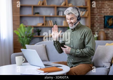 Smiling man wearing a headset and waving, participating in a video call on a laptop from his home office, highlighting remote work and virtual communication Stock Photo
