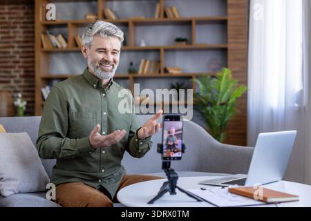 Smiling mature man recording a video lesson for an online audience, sitting comfortably on a gray sofa in a home office, sharing knowledge through digital media Stock Photo