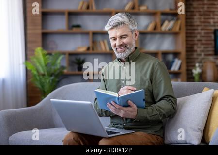 Mature man smiling, sitting comfortably on a sofa, holding a notebook and writing while working from a laptop, capturing the essence of flexible remote work Stock Photo