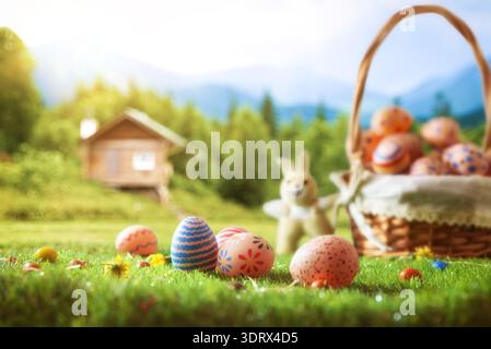 Decorated Easter eggs in a basket on green grass with a small bunny toy. Sunny spring landscape with wooden cabin and mountains blurred in background. Stock Photo