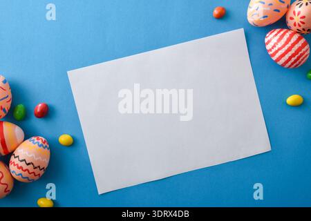 Top view of a blank white card surrounded by colorful painted Easter eggs and candy on a bright blue background, perfect for holiday greeting or invit Stock Photo