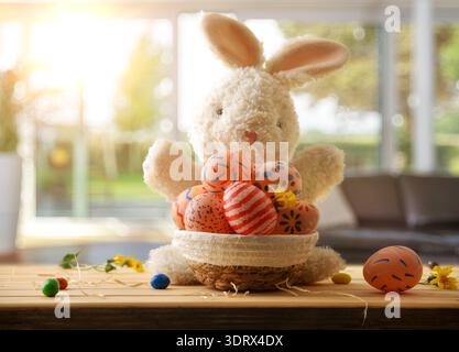 A fluffy plush bunny holding a basket of colorful hand-painted Easter eggs on a wooden table, with warm sunlight and a cozy indoor spring background. Stock Photo