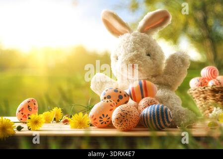 Fluffy toy bunny with painted Easter eggs and yellow flowers on a wooden table, sunny spring garden background with soft bokeh and warm light. Stock Photo