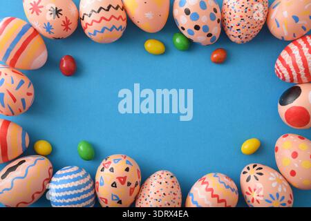 Colorful hand-painted Easter eggs forming a decorative frame on a bright blue background with scattered candy, leaving space in the center for text. Stock Photo