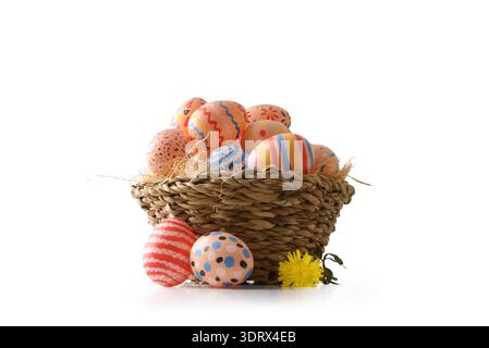 Rustic woven basket filled with brightly painted Easter eggs in striped and dotted patterns, placed on a white background with a yellow spring flower. Stock Photo