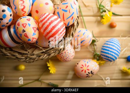 Close-up of pastel Easter eggs with vibrant hand-painted patterns in a wicker basket on a wooden table, decorated with yellow spring flowers. Stock Photo