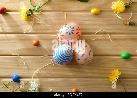 Top view of three pastel Easter eggs with floral and striped patterns on a wooden surface, surrounded by spring flowers and colorful candies. Stock Photo