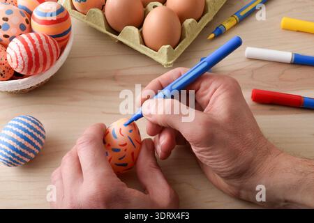 Close-up of adult hands painting a brown Easter egg with a blue marker on a wooden table. Decorated eggs, carton of eggs and colorful pens create a fe Stock Photo