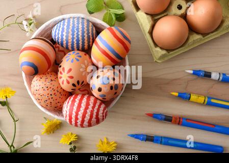 Top view of vibrant hand painted Easter eggs in a white bowl on a wooden table. Egg carton, watercolor pens and yellow flowers enhance the cheerful sp Stock Photo