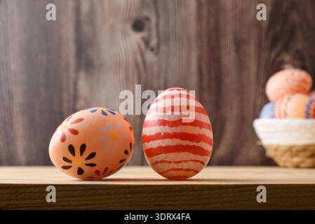 Two hand painted Easter eggs with floral and striped patterns on a wooden shelf, with a blurred basket of eggs in the background. Stock Photo