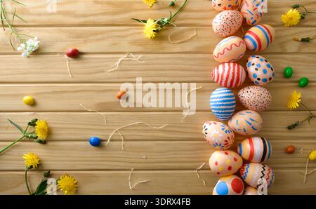Top view of hand-painted Easter eggs arranged as a border on a wooden background with spring flowers and colorful candies, leaving copy space. Stock Photo