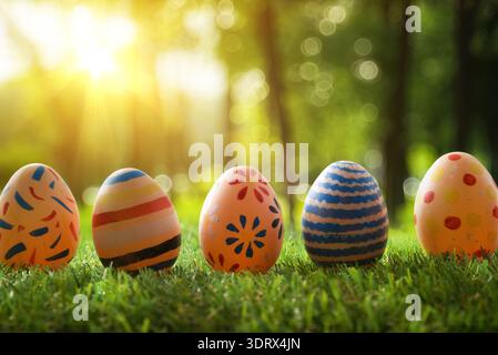 Five colorful Easter eggs lined on green grass with warm sunlight and forest bokeh background, vibrant spring celebration scene. Stock Photo