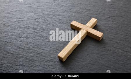 Simple wooden cross placed on a dark textured stone surface with soft natural light. Minimal Christian symbol concept with copy space. Stock Photo