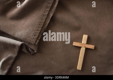 Small wooden cross resting on brown fabric with soft folds. Warm, rustic composition representing Christianity, faith, prayer and spiritual reflection Stock Photo