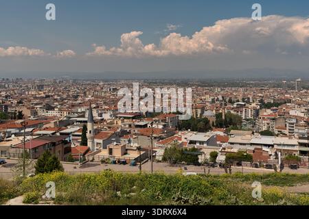 Panoramic cityscape of Manisa, Turkey, with dense buildings, red roofs, minarets, and distant mountains under a partly cloudy sky Stock Photo