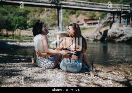 Two friends enjoy a sunny riverside moment by the shore with drinks Stock Photo