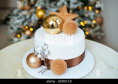 A round cake sits on a white plate. It has gold ornaments and other decorations on top. A Christmas tree with lights is in the background. The scene i Stock Photo