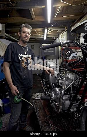 Mechanic in a workshop setting, standing next to a partially assembled custom chopper-style motorcycle. Stock Photo