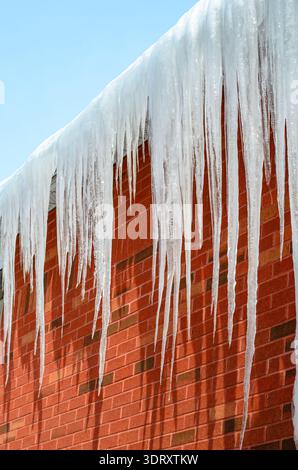 Brick building with long icicles hanging below a roof on a winter day, with a clear blue sky above. Stock Photo