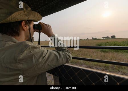 A local safari guide standing on top of his vehicle looking through binoculars searing for wildlife to show the tourists, Nairobi National Park, Kenya Stock Photo