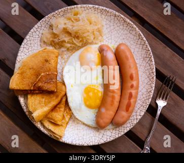 Fried sausages with scrambled eggs, pancakes and cabbage on a wooden background Stock Photo