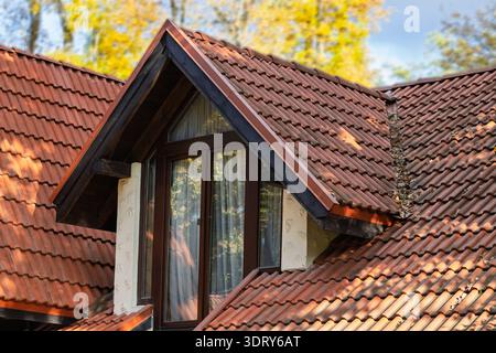 The warm hues of autumn foliage are reflected in the glass of a dark-framed dormer window set within a classic terracotta tiled roof structure Stock Photo