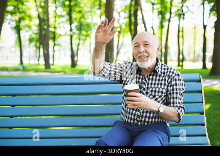 Elderly Man Enjoys Coffee While Sitting in Park and Waves at Passersby During Sunny Day Stock Photo