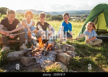 Five kids group Boys and girls cheerfully laughed and roasted sausages on sticks over a campfire flame near the green tent. Outdoor active time spendi Stock Photo
