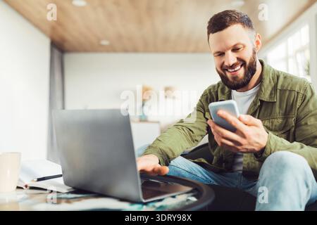 Male Entrepreneur Using Smartphone And Laptop Working Online At Home Stock Photo