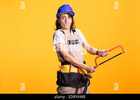 Serious white woman in construction gear uses handsaw, portraying home improvement or labor effort. Female contractor stands in safety helmet and tool apron against isolated yellow background. Stock Photo