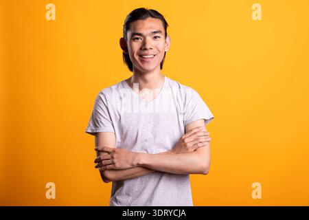 Smiling young Asian man stands confidently with folded arms, showing happiness and fulfillment. Proud and satisfied male model poses, radiating positive attitude and cheerful self assurance. Stock Photo