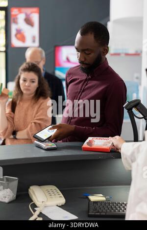 African american man taps mobile device on pos terminal to complete pharmacy purchase while other clients wait in line. Black male adult uses cellphone at retail medical store to pay for medicine. Stock Photo