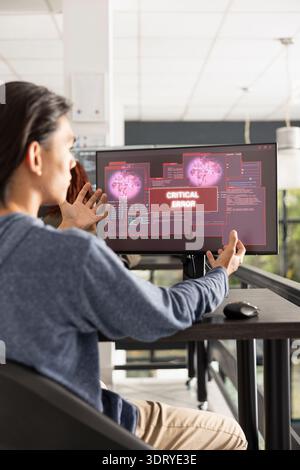 Portrait of male programmer gesturing at alarming error alert on desktop monitor. Asian software developer sits at desk in modern office, reacting to critical error message on computer screen. Stock Photo
