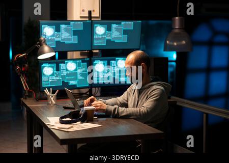 Bearded white man works late night at office laptop, writing code and debugging software. Caucasian male programmer focuses on cybersecurity, cloud computing and preventing cybercrime online. Stock Photo