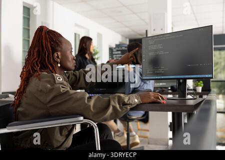 Black woman programmer focuses on coding at office desk with desktop PC and monitor. Confident IT professional in wheelchair showcases diversity, accessibility and ambition in tech careers. Stock Photo