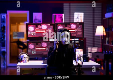 Masked cybercriminal doing espionage campaign from secret base of operations. Rogue software developer wearing anonymous mask using smartphone to record demands from secret underground lair Stock Photo