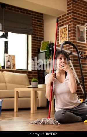 Caucasian woman sits on floor with mopping stick, yawning and covering her mouth, exhausted after long day of housecleaning. Young lady takes moment to rest before completing household chores. Stock Photo