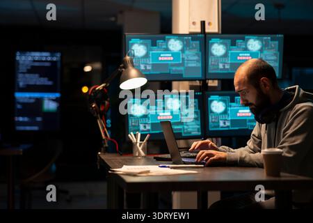 Dedicated male programmer analyzing system data and writing secure code on laptop. Concentrated developer enhancing cloud computing protection and addressing cybercrime risks during night shift. Stock Photo
