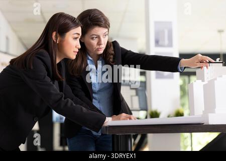 Focused diverse women analyze blueprints and scale model, preparing for upcoming construction site visit. Female architects making necessary adjustments to ensure building project meets requirements. Stock Photo