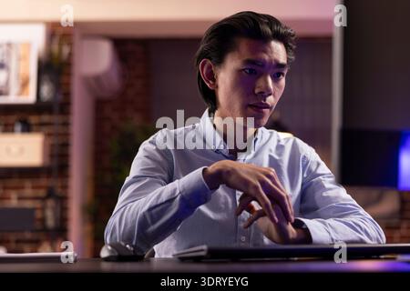 Asian remote worker sits at desk during evening hours, reading emails and reviewing business data on desktop PC. Focused male entrepreneur researching strategies to improve small business plans. Stock Photo