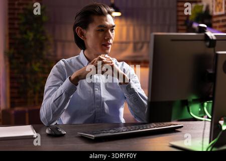 Serious young asian man watching online course on computer, learning financial strategies from virtual class. Focused male entrepreneur participates in digital education session from cozy apartment. Stock Photo