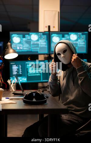 Anonymous man adjusting mask and hoodie while sitting at desk surrounded by glowing system monitors. Programmer with concealed identity seated in dark office room, planning to steal sensitive data. Stock Photo