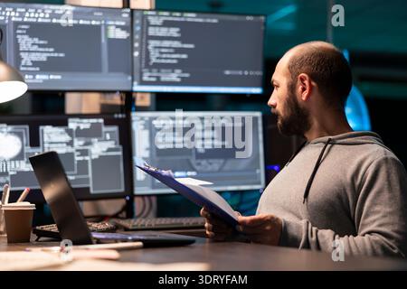 Programmer examines paperwork with serious expression and compares notes to code displayed on surrounding monitors. Developer with clipboard glancing at large displays of data, ensuring accuracy. Stock Photo