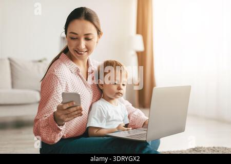 Woman using laptop and smartphone, child boy sitting on mother's knees and watching cartoons online on computer Stock Photo