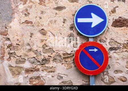 Blue directional arrow sign and red no parking sign mounted on a rustic weathered stone wall in a historic european village featuring textured masonry Stock Photo